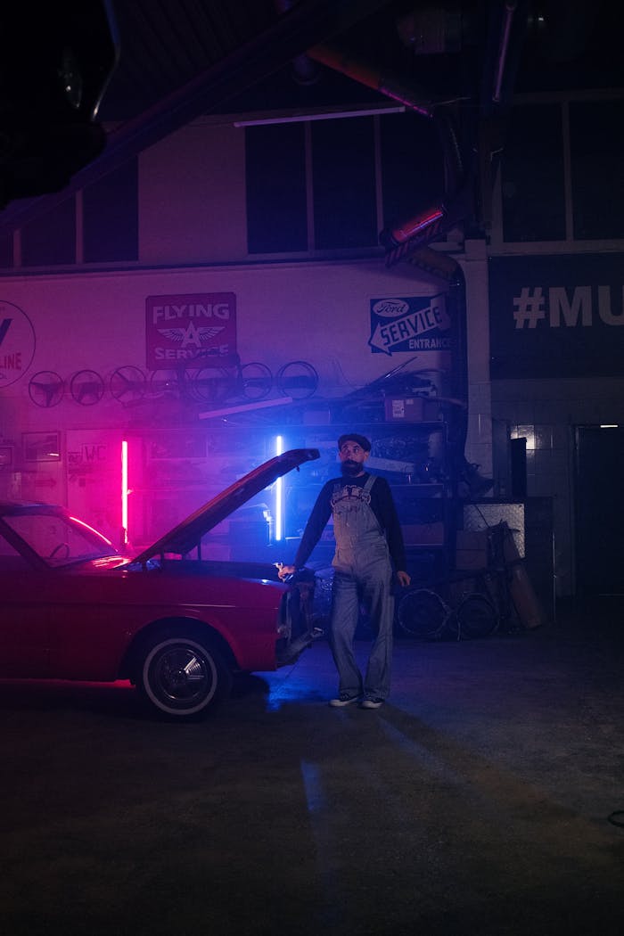 Mechanic working on a vintage car under neon lights in a dim garage setting.